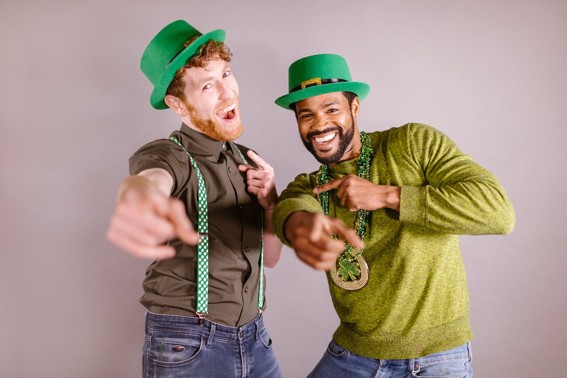 Two man employees in a green leprechaun hat, one in green suspenders and one in clover necklace for outfit ideas as a St Patrick's Day office ideas.